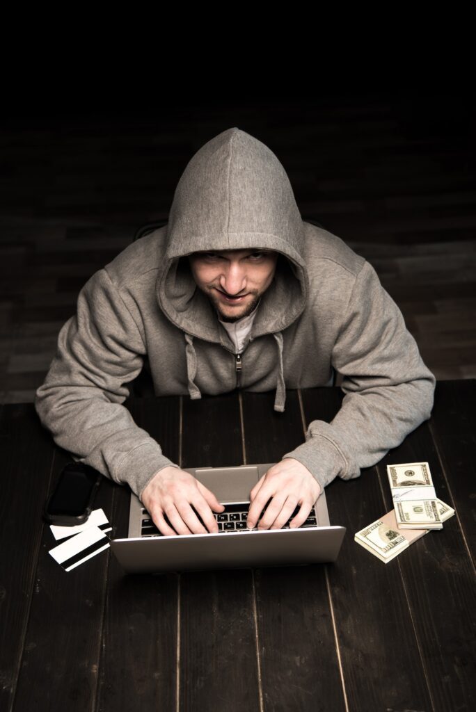 Young hacker in hoodie sitting at table and using laptop